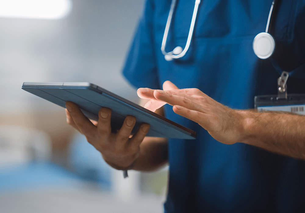 Person's torso showing in photo, covered in blue hospital scrubs, wearing a stethoscope around the neck and holding a tablet device