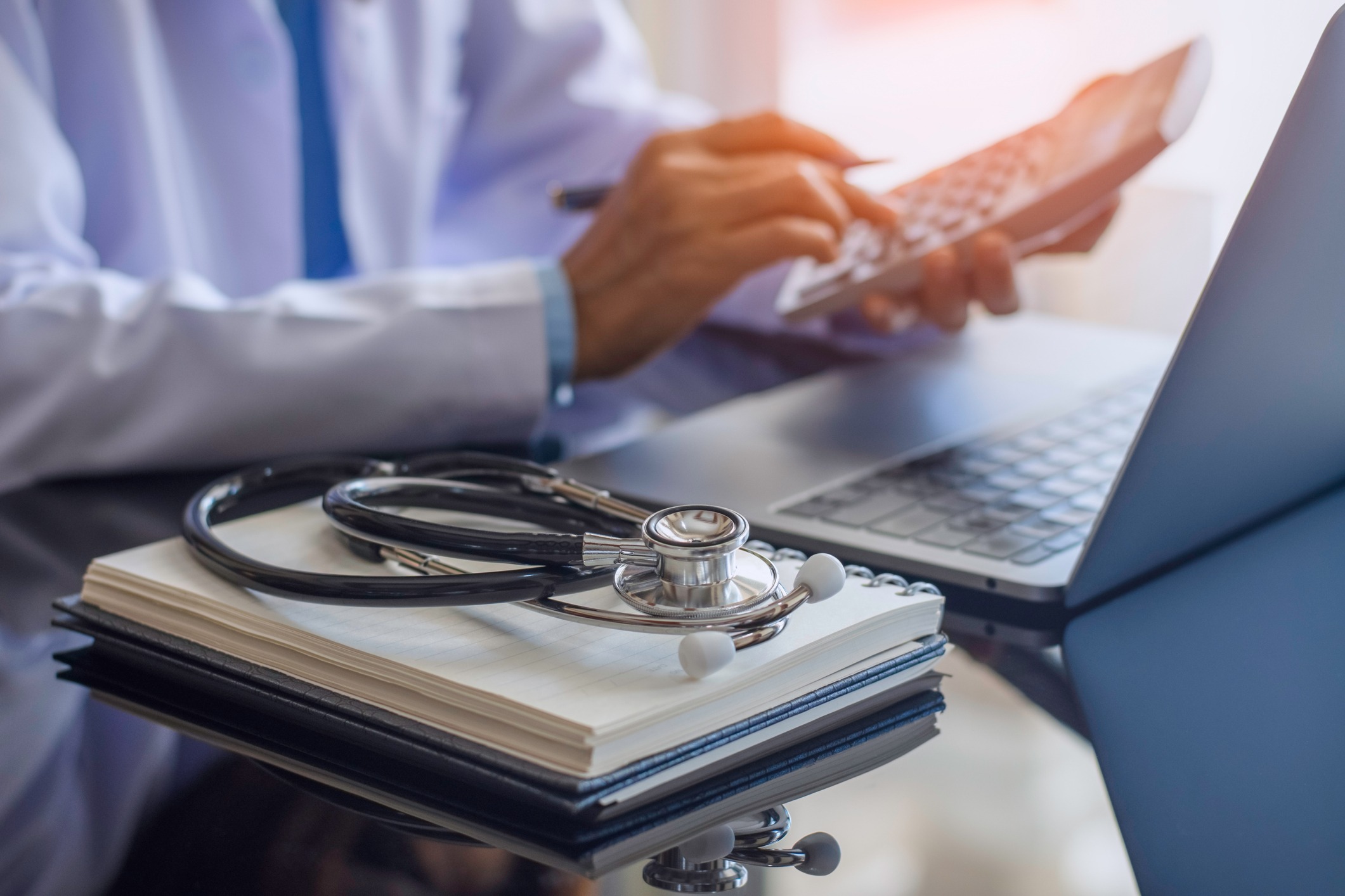 Female doctor or practitioner using calculator and work on laptop computer with medical stethoscope and notebookon the desk at clinic or hospital. Medical healthcare costs ,fees and revenue concept