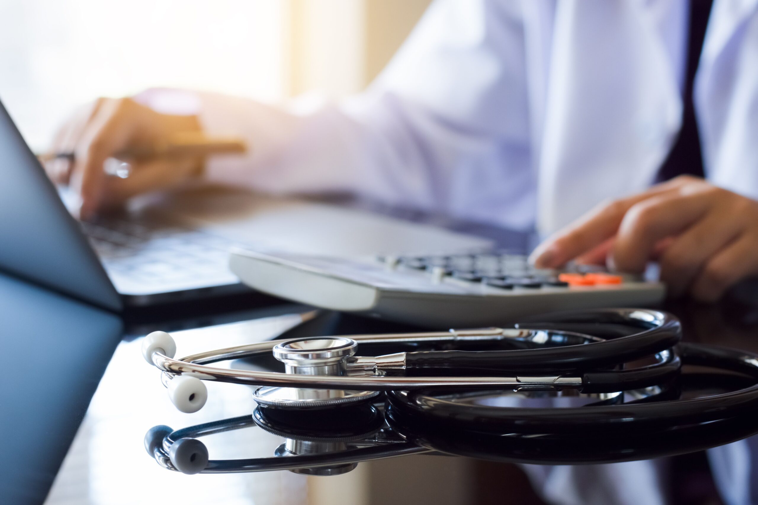 A person sat at a lap top while using a calculator with a stethoscope next to them - representing health costs