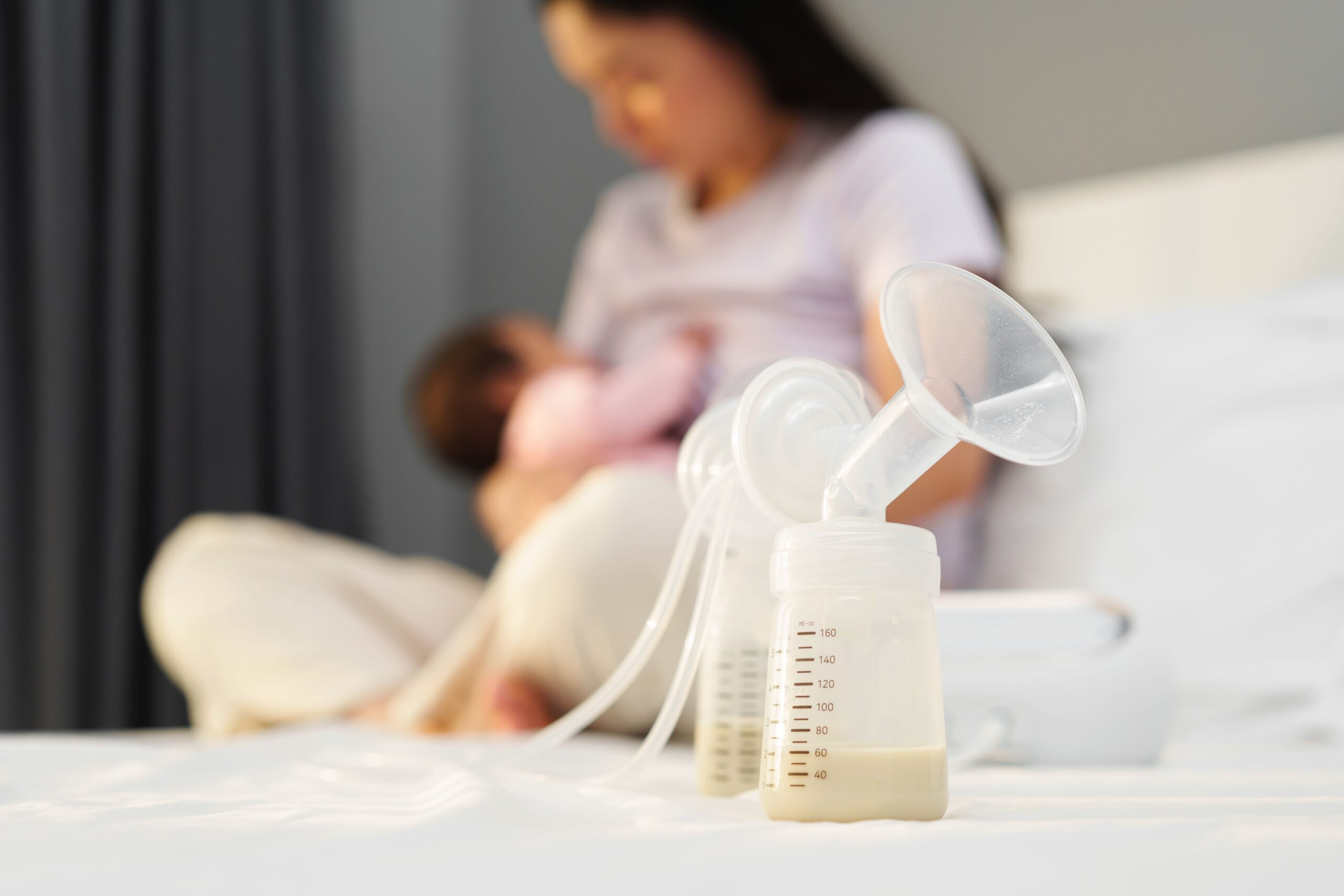 Breast milk in bottles of electric breast pumping set on a bed with mother breastfeeding newborm baby in the background